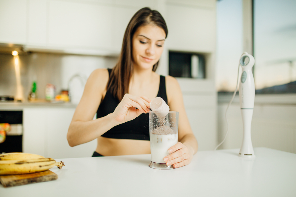 Woman with immersion blender making banana chocolate protein with colostrum powder milkshake smoothie.Adding a scoop of low carb whey protein mix to shake after a home workout.Diet after the gym.Healthy lifestyle