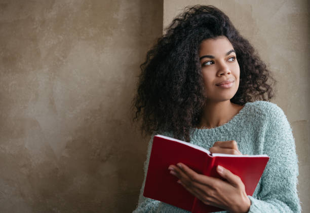 Young beautiful African American woman holding red book, looking at window and smiling. University student studying, learning language, sitting at library. Portrait of young pensive writer taking notes, woman reading a book, empowering female reading books